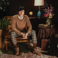 Man in a brown sweater and plaid pants sitting in an armchair.